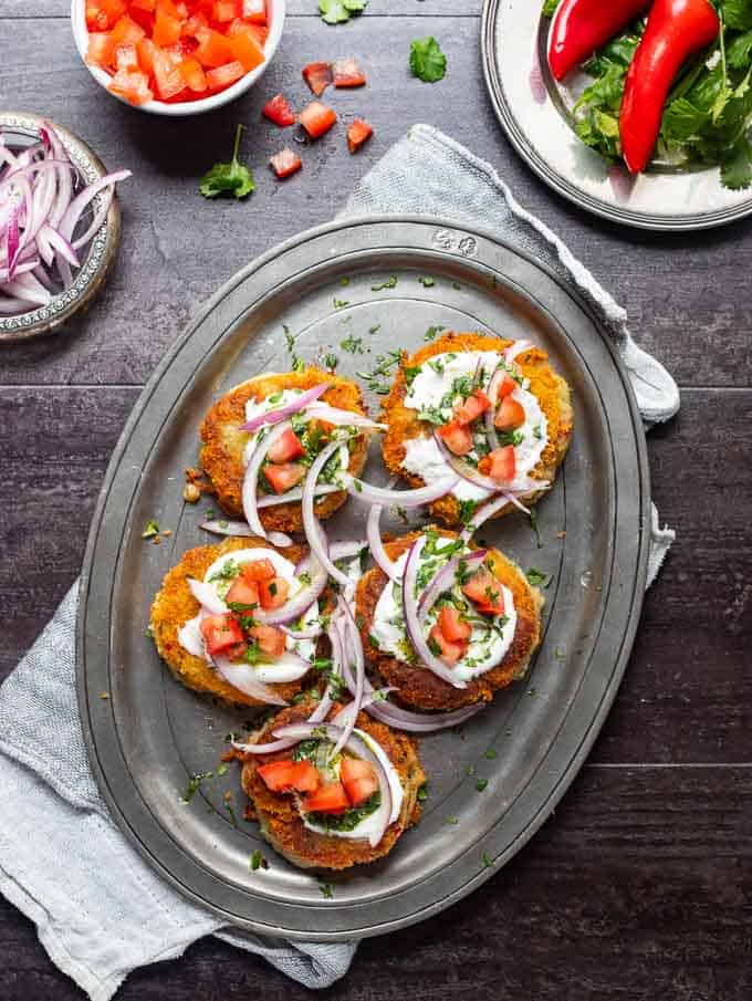 Aloo tikki's on a metal platter and garnished with tomatoes, onions, yogurt, mint chutney and cilantro.