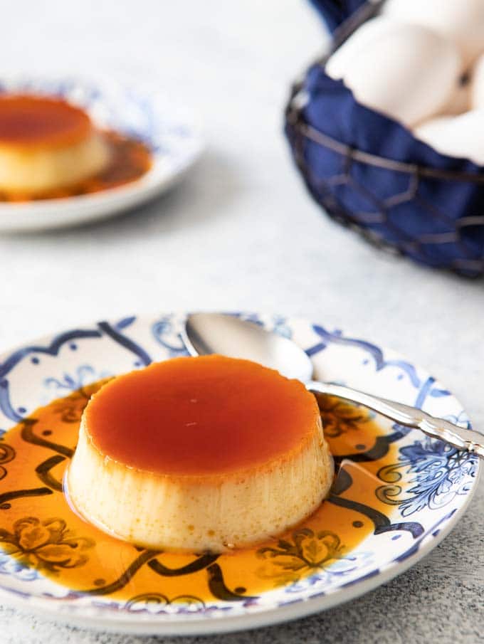 Single serving of Spanish flan on a plate with a basket of eggs in the background.
