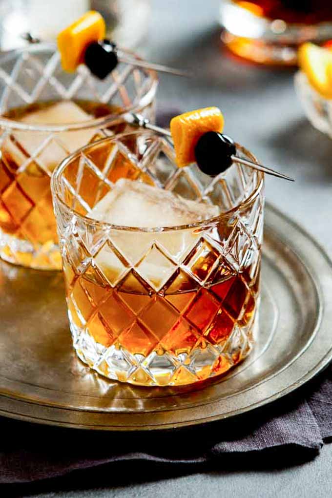 Two rocks glasses on a tray with a bourbon manhattan and a cherry and orange peel garnish.