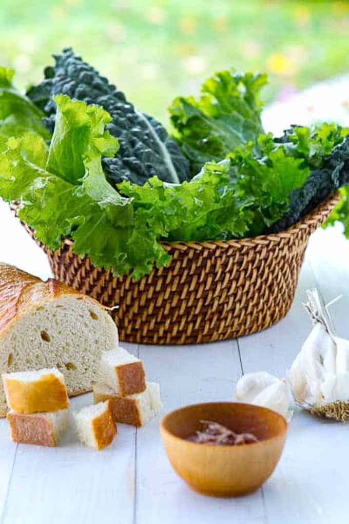Basket with spicy greens including mustard greens and kale, with a loaf of bread, anchovies and garlic in front.
