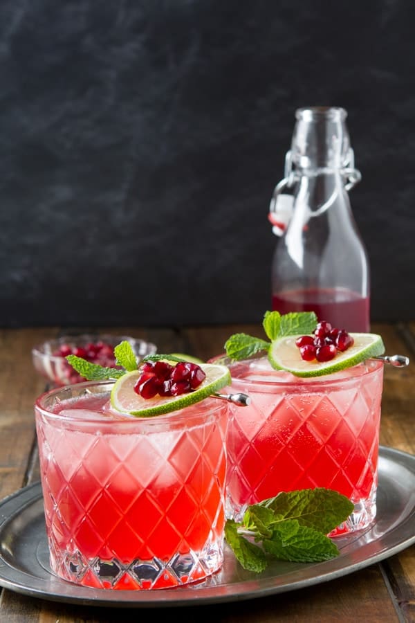 Two glasses of a Gin fizz cocktail flavored with pomegranate with a bottle of grenadine in the background.
