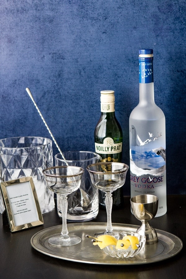 A bar set up for making vodka martini's - vodka, vermouth, glasses, a mixing glass and spoon, jigger, ice bucket and cocktail garnish on a platter.
