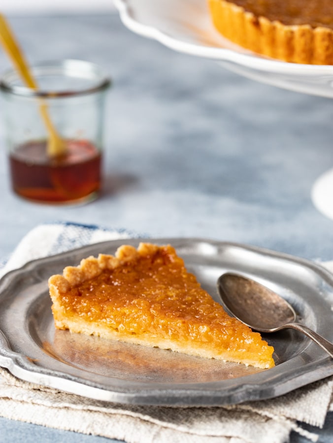 A single slice of treacle tart on a plate with golden syrup in the background.