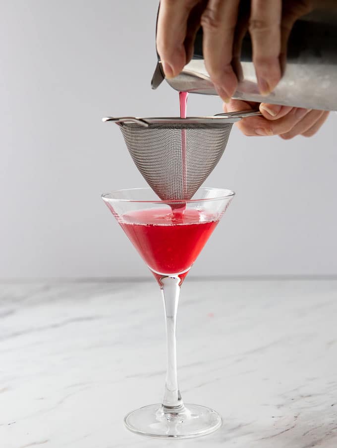 double straining vodka and cranberry drink into a cocktail glass.