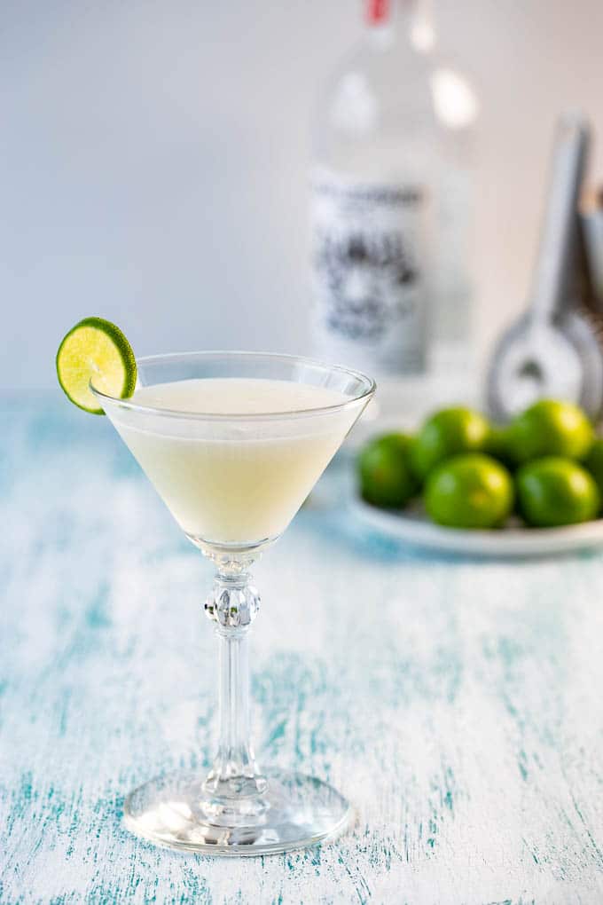 A key lime martini in a martini glass with a bottle of vodka and a dish of limes in the background.