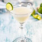A single glass of a key lime martini with a bottle of vodka and limes in the background.