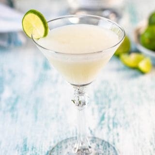 A single glass of a key lime martini with a bottle of vodka and limes in the background.