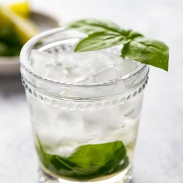 Single glass of basil and lime vermouth cocktail with a basil leaf garnish and limes in the background.