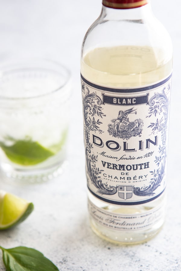 Bottle of Dolin blanc vermouth with a glass in the background with lime and basil. 
