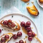 Grilled red grapes on top of whipped ricotta corstini on a plate with two skewers of grilled red grapes.