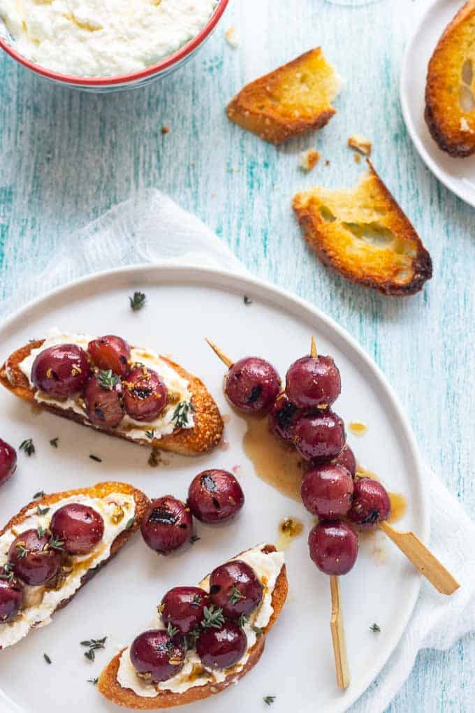 Whipped ricotta crostini on a plate with grilled grapes and a bowl of whipped ricotta in the background. 