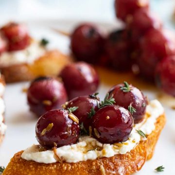 Close up of grilled red grapes on top of whipped ricotta, sprinkled with fresh thyme on a large white plate.