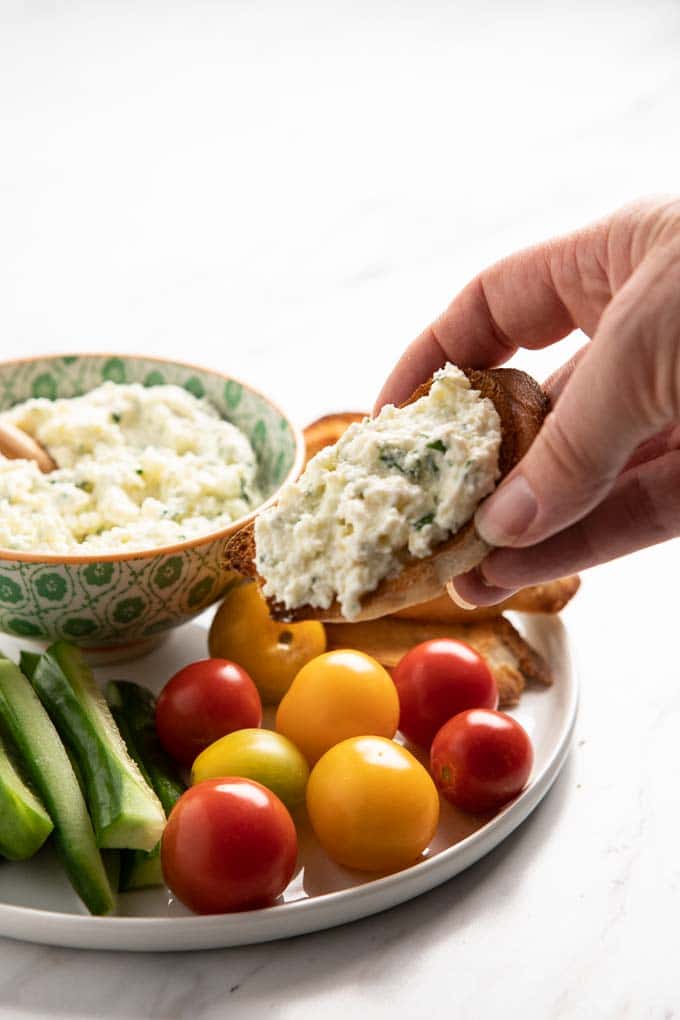 Bowl of whipped ricotta spread on a baguette slice with a bowl of whipped ricotta and vegetables in the background.