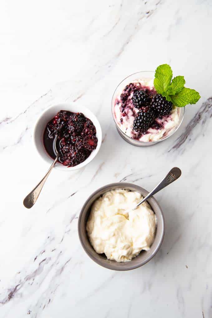 Overhead photo of a simple  blackberry fool recipe in a glass with a bowl of fresh blackberries and fresh whipped cream on the side. 