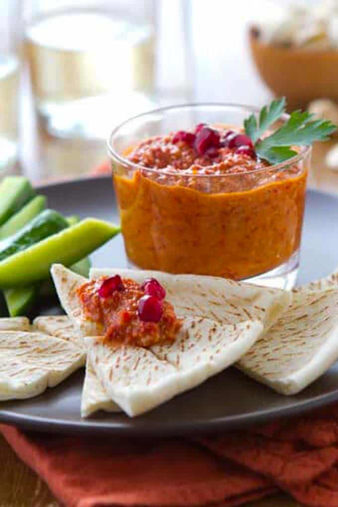 A small bowl of muhammara dip on a plate with  pita bread  and  cucumber.
