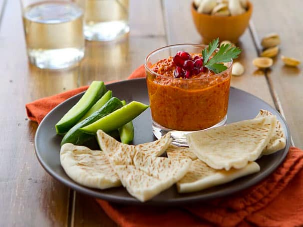 Muhammara dip recipe in a bowl with pita and cucumber slices. 
