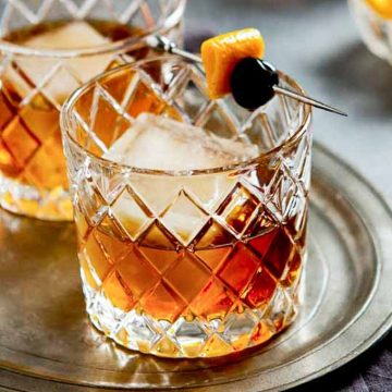 Two rocks glasses on a tray with a bourbon manhattan and a cherry and orange peel garnish.