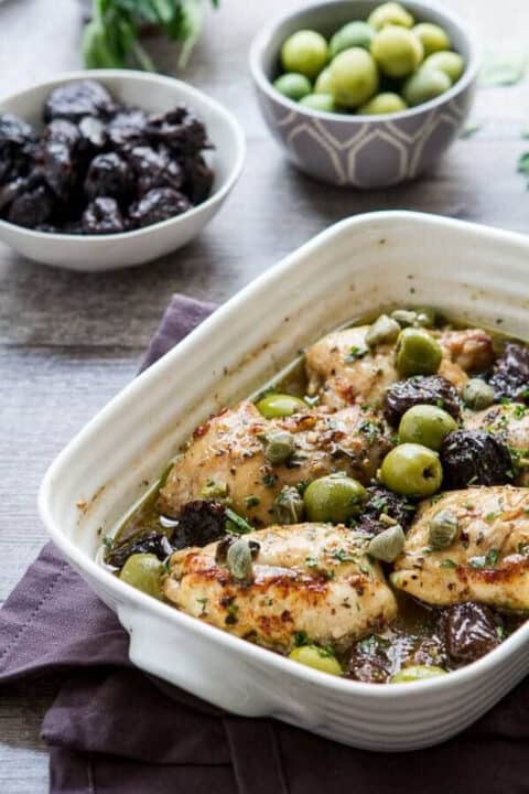 Chicken Marbella recipe in a white casserole dish with prunes and olives in the background.